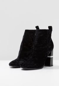 Schwarze Samt-Stiefeletten mit spitzem Zeh und blockiertem Absatz, die mit einem silbernen Akzentband versehen sind. Die Textur ist weich und samtig.