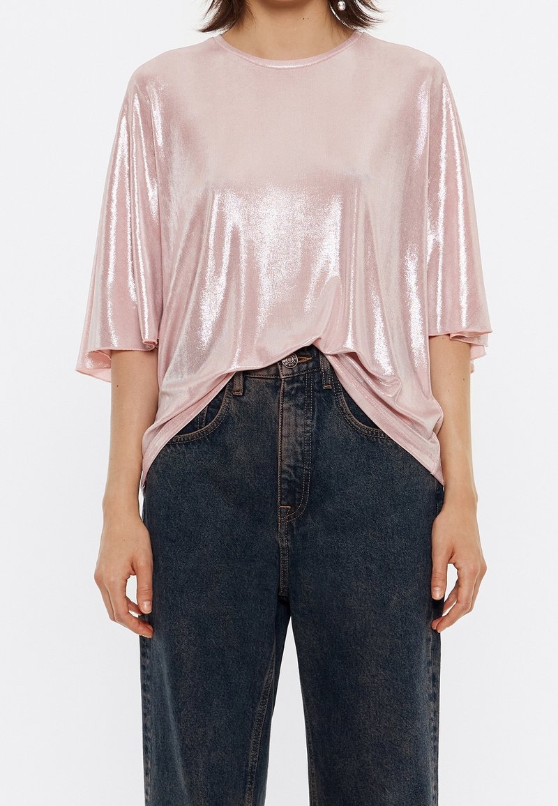 Osoba ubrana w błyszczącą różową luźną bluzkę lekko włożoną do wysokich, ciemnoniebieskich dżinsów, stojąca na jednolitym tle.