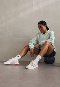 Hvide og lyserøde træningssko med mesh- og gummidetaljer, parret med hvide sokker og en mintgrøn sweatshirt, der ligger på et gråt gulv.