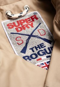 Tessuto beige con una toppa cucita che mostra "SUPER DRY" in rosso e "THE ROGUE" in blu, con fucili incrociati e un accento della Union Jack.