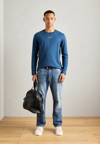 Man in een blauwe lange mouwen shirt, vervaagde blauwe spijkerbroek, witte sneakers, met een zwart leren tas, staand op een houten vloer tegen een effen muur.