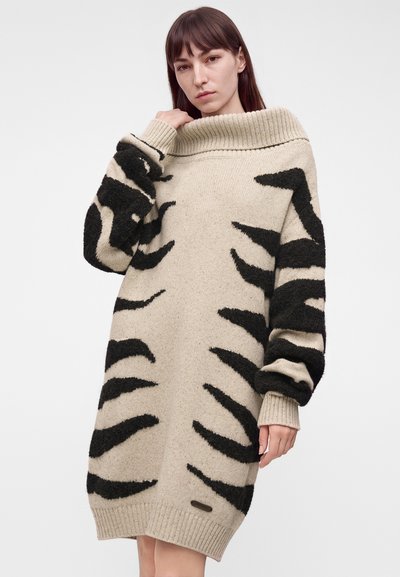 Νεαρή γυναίκα που φοράει μια μπεζ oversized φορεματόπλεκτο με μαύρες αφηρημένες ρίγες τίγρης και φαρδιά διπλωμένη γιακά, στέκεται μπροστά σε απλό φόντο.