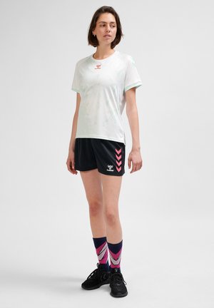 Witte sportshirt met mintgroene accenten, gecombineerd met zwarte shorts met roze chevron-details, en gestreepte roze en marineblauwe sokken.