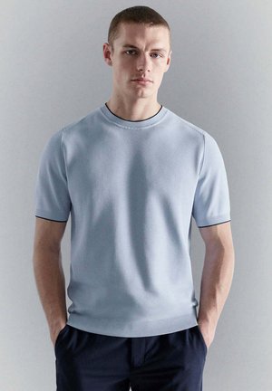 Jonge man met kort haar die een lichtblauw, gebreid T-shirt met korte mouwen en een donkerdere broek draagt, staand tegen een effen lichtgrijze achtergrond.