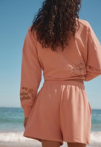 Felpa color pesca con spalle scese e dettagli ricamati, abbinata a pantaloncini coordinati con vita elastica e tasche laterali.