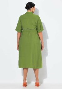 Abito verde, a metà coscia, con maniche corte, caratterizzato da una vita con cintura e una consistenza morbida. Indossato con sandali arancioni con tacco.