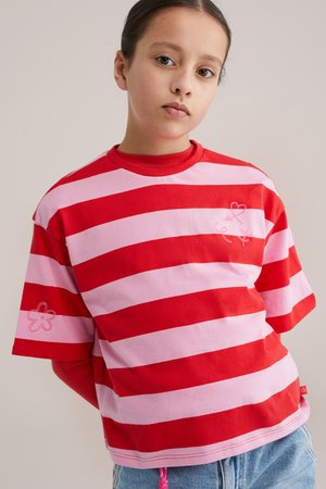 Persoană tânără purtând o cămașă cu dungi roșii și roz și modele florale, stând cu brațele pe spate, expresie neutră, fundal bej.