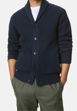 Homme portant un cardigan en tricot bleu marine boutonné sur une chemise blanche, les mains dans les poches d’un pantalon vert olive, cadré au niveau du cou et des cuisses.