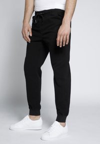 Pantaloni da jogging neri con vita elastica e cordino. Polsini alle caviglie e tasche laterali. Indossati con sneakers bianche.