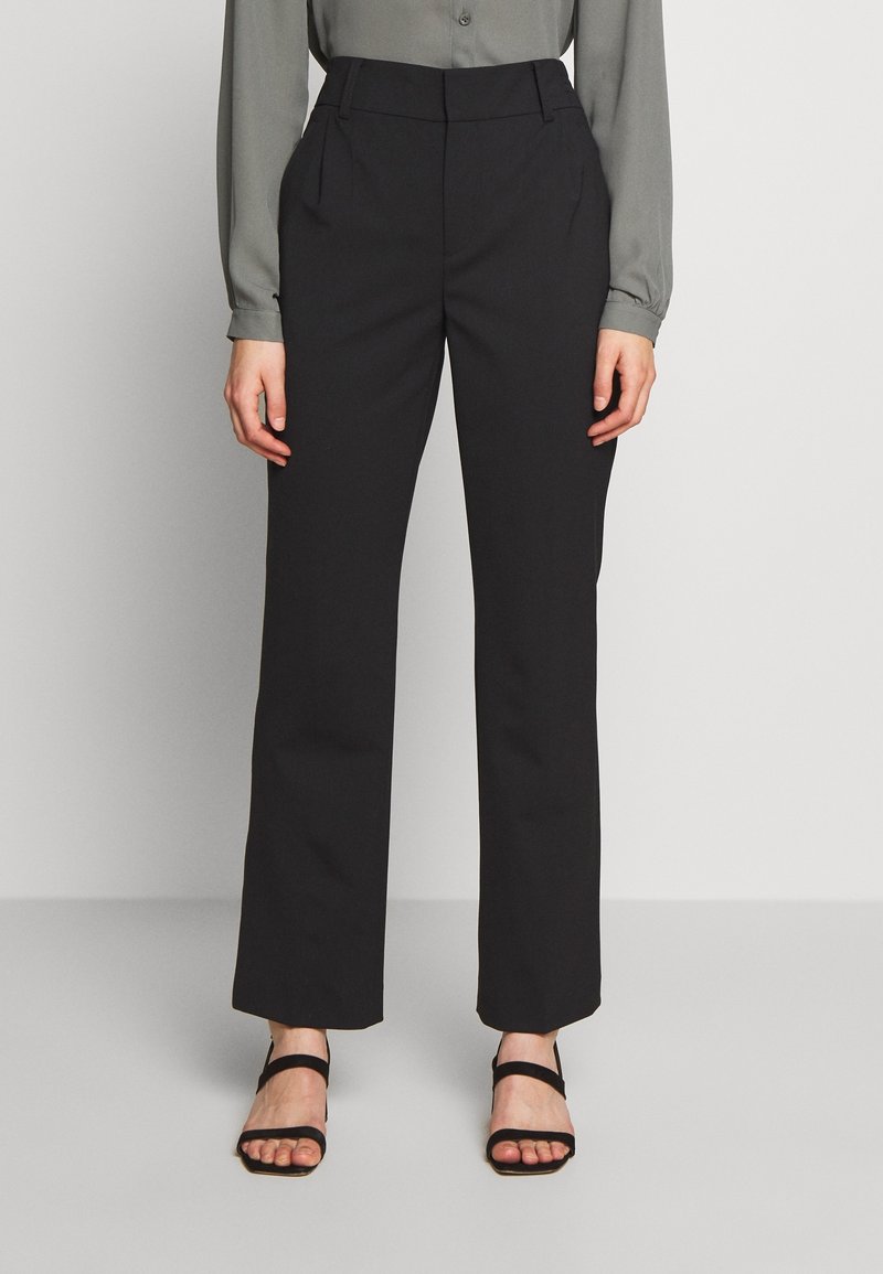 Femme portant un pantalon noir droit, une chemise grise à manches longues et des sandales noires à bouts ouverts, debout devant un fond blanc uni.