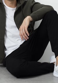 Sudadera con cremallera verde oscuro sobre una camiseta blanca, combinada con pantalones negros y zapatillas blancas, sentado en un suelo gris.