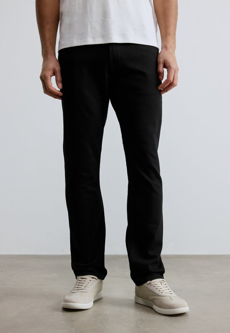 Μαύρα τζιν σε στενή γραμμή, κατασκευασμένα από τζιν ύφασμα, με λείο υλικό. Φορεμένα με μπεζ αθλητικά παπούτσια, στέκονται μπροστά σε ουδέτερο φόντο.