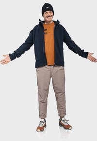 Marineblaues Fleece-Jackett mit Kapuze, orangefarbenes T-Shirt mit Aufdruck, beige Hose und mehrfarbige Sportschuhe. Modell steht mit offenen Armen.