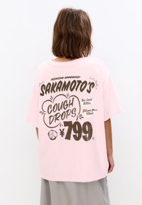 Lyserød bomulds t-shirt med løs pasform, featuring store sorte bogstaver på ryggen, der står "Sakamoto's Cough Drops" og et prisskilt på ¥799.