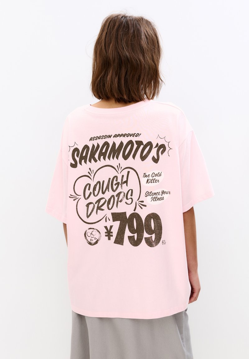 Lyserød bomulds t-shirt med løs pasform, featuring store sorte bogstaver på ryggen, der står "Sakamoto's Cough Drops" og et prisskilt på ¥799.