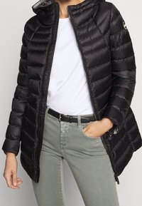 Černá prošívaná bunda s kapucí, s prošitými částmi, zipem a bočními kapsami, doplněná bílým tričkem a zelenými džíny.