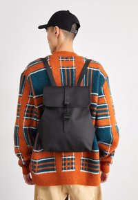 Zaino nero con chiusura a patta, spallacci regolabili e una fibbia. Indossato sopra un maglione a fantasia arancione, verde e bianco.
