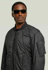 Giacca bomber nera con chiusura frontale a zip, colletto a costine e tessuto strutturato; presenta una tasca a toppa sulla manica e occhiali da sole lisci.