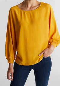 Blouse jaune légère avec un col arrondi, des manches longues ballon avec des poignets élastiques, et une coupe décontractée, associée à un jean foncé.