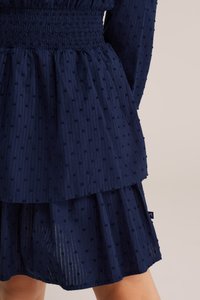 Tengerészkék ruha smockolt derékrésszel, réteges fodros szoknyával és texturált anyaggal, melyen apró kiemelkedő pöttyök találhatók. Oldalsó címke látható.