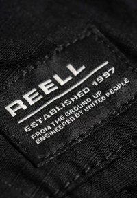 Schwarzes Stoffetikett mit "REELL" in fettem weißen Text, sowie "GEGRÜNDET 1997" und zusätzlichen Informationen über die Ursprünge und die Technik der Marke.