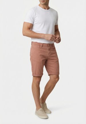 Uomo che indossa pantaloncini rosa polvere arrotolati, maglietta bianca e scarpe da ginnastica beige, in piedi contro uno sfondo bianco.