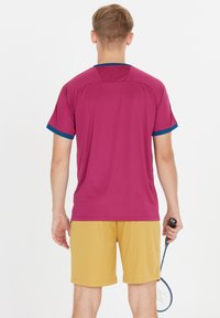 Rød sports t-shirt med blå detaljer, parret med gule shorts. Stoffet ser letvægtsagtigt ud, velegnet til atletiske aktiviteter.