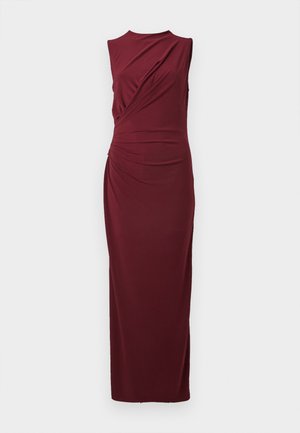 Beigių fono fone, be rankovių, grindų ilgio bordo suknelė su sutraukta medžiaga priekyje ir prigludusia siluetu.