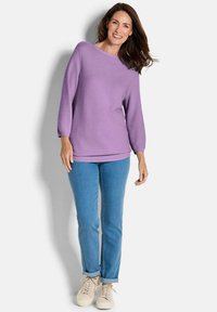 Lavendelfarbener Pullover mit langen Ärmeln und Bootsausschnitt, kombiniert mit hellblauen Jeans und beigen Sneakers. Weicher Stoff, lässige Passform.