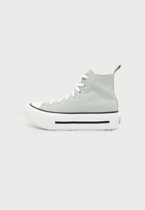 Zapatilla de lona gris clara de caña alta con cordones blancos, suela gruesa de goma blanca y detalle de raya negra sobre un fondo liso.