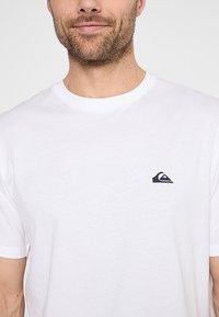 Camiseta de algodón blanca con cuello redondo y mangas cortas, con un pequeño logo en color marino en la parte izquierda del pecho. Textura suave, diseño casual.