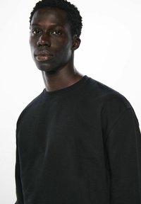 Schwarzer Baumwollpullover mit Rundhalsausschnitt, langen Ärmeln und strukturiertem Stoff. Eng anliegend an Schultern und Brust. Schlichtes Design.