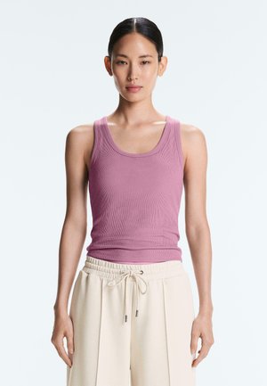 Jeune femme aux cheveux lisses et foncés attachés, portant un débardeur mauve côtelé sans manches et un pantalon beige à cordon, debout devant un fond uni.