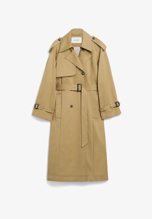 Gabardina beige de doble botonadura con cinturón, hombreras y correas en los puños, llevada sobre un jersey de cuello alto negro y botas negras hasta la rodilla.