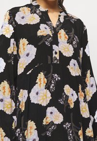 Zwarte lange mouw jurk met gele en witte bloemmotieven en een gedeeltelijke knoopsluiting aan de voorkant van de kraag.
