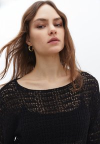 Svart stickad topp med en texturerad, öppen vävningsmönster, som har en vid halsringning och långa ärmar. Modellen visar naturlig sminkning och accessoarer.