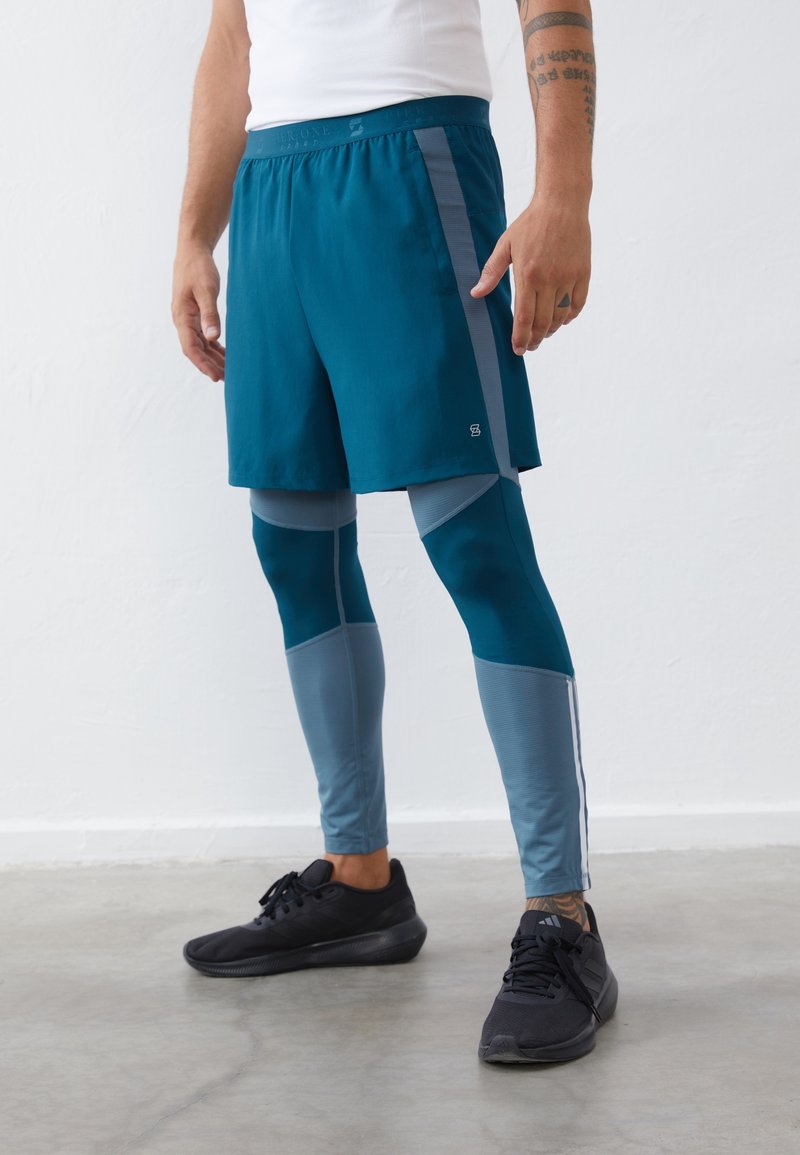 Shorts de sport turquoise avec des panneaux latéraux gris, associés à des leggings teal présentant des accents en mesh et des rayures. Des baskets noires complètent le look.