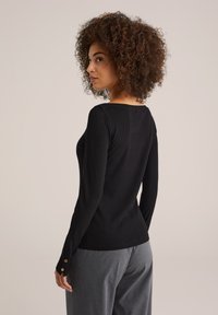Zwarte ribbelige lange mouw top met een brede halslijn, voorzien van knoopaccenten bij de manchetten, gecombineerd met grijze gestreepte broeken.