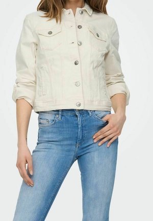 Vrouw die een crèmekleurig spijkerjack met knopen en opgerolde mouwen draagt, en lichtblauwe slimfit jeans, hand rustend op de heup.
