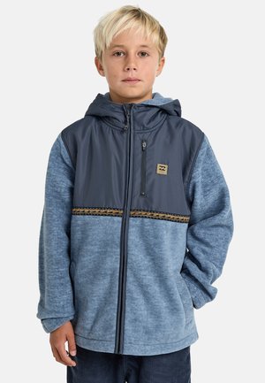 Blond boy wearing a blue zip-up hooded jacket with a dark upper section and patterned trim, standing with one hand in pocket.