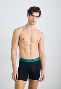 Boxer aderenti neri con una fascia verde di Calvin Klein, caratterizzati da una texture liscia e un design aderente, indossati da un modello maschile.