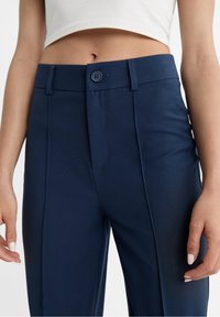 Pantalon bleu marine avec une texture lisse, doté d'une fermeture à bouton à l'avant, de passants de ceinture et de plis nets pour une coupe ajustée.