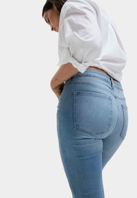 Úzké džíny v světle modrém denimovém odstínu s vysokým pasem. Obsahují klasické zadní kapsy a hladkou texturu. Jsou kombinovány s bílou blůzou s ohrnutými rukávy.