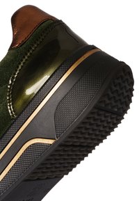 Groene en metalen koperen sneakerhak met zwarte, getextureerde rubberen zool en gouden accentstreep, schuin geplaatst om het zoolpatroon en de hakedetails te tonen.