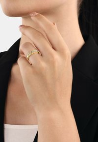 Złoty pierścionek z rzędem przezroczystych kamieni wzdłuż obrączki, noszony na dłoni z neutralnym manicure, na tle ciemnej kurtki.