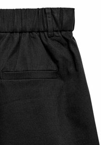 Gros plan sur un tissu noir montrant une taille élastique, une passants de ceinture, et un détail de poche arrière horizontale unique sur un vêtement.