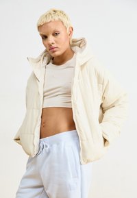 Casaco acolchoado bege claro com capuz, apresentando um fecho na frente, usado sobre um top cropped bege e calças azul claro. Textura suave, design minimalista.