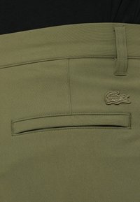 Pantalone od tkanine u maslinasto zelenoj boji s stražnjim džepom na kojem je izvezeni logo. Glatka tekstura, standardni kroj i čist dizajn.