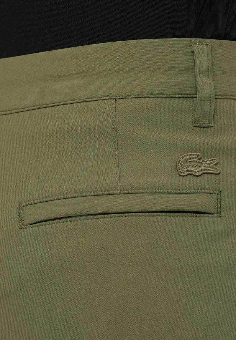 Pantaloni in tessuto verde oliva con una tasca posteriore decorata con un logo ricamato. Texture liscia, vestibilità standard e design pulito.