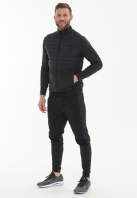 Sort atletisk jakke med lynlås fortil, polstret panel på brystet og tætsiddende ærmer. Sammen med sorte tapered joggers og grå sko.